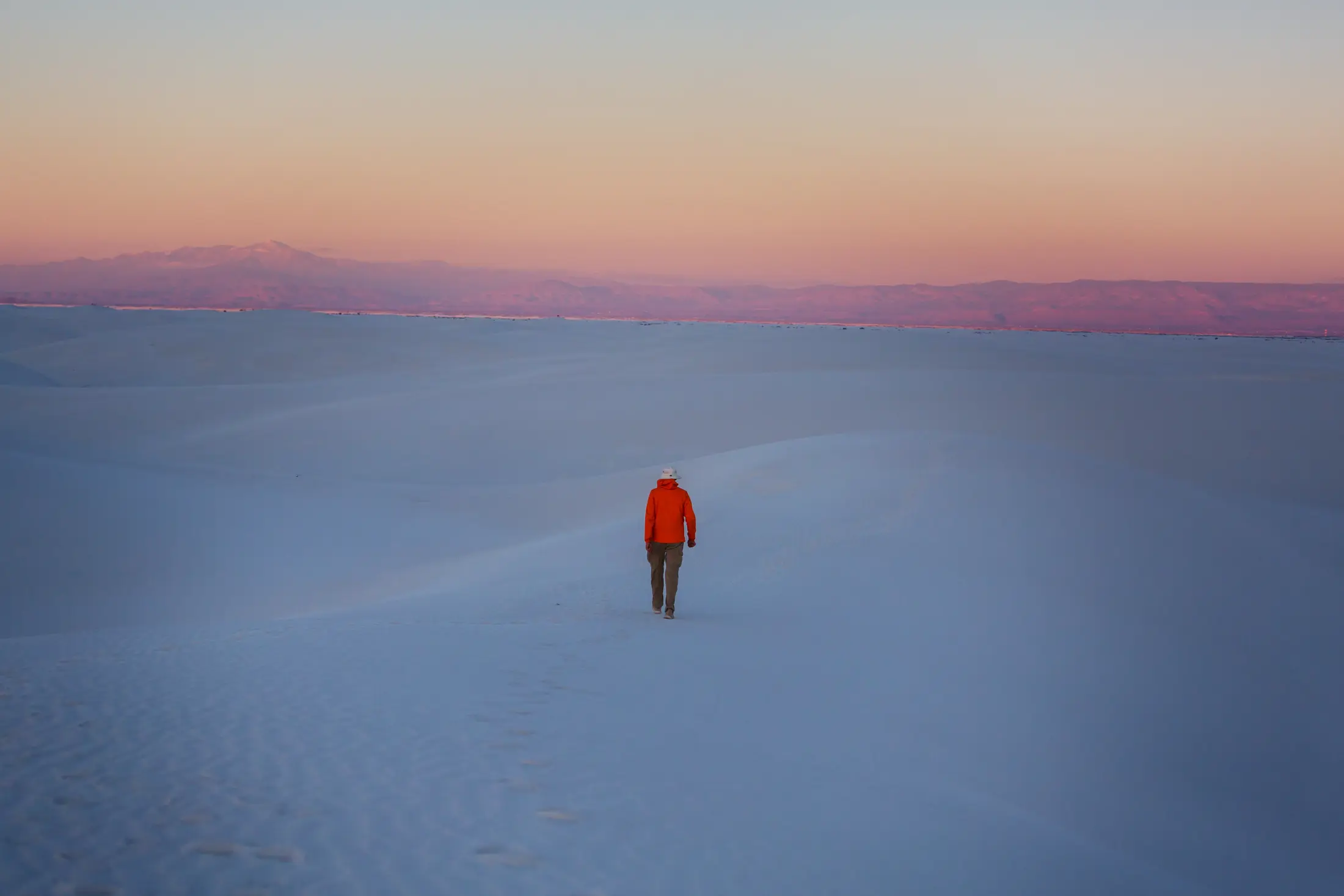 A lone figure in a red jacket walking across white dunes at sunrise